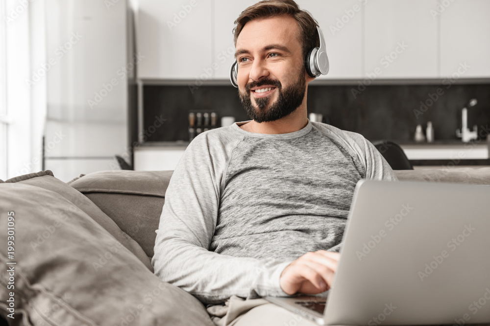 Attractive guy 30s in casual clothing sitting on couch, and listening to music on laptop wearing earphones Attractive guy 30s in casual clothing sitting on couch, and listening to music on laptop wearing earphones