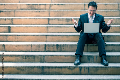 Happy Asian businessman working on laptop while sitting on the city stairs. Copy space