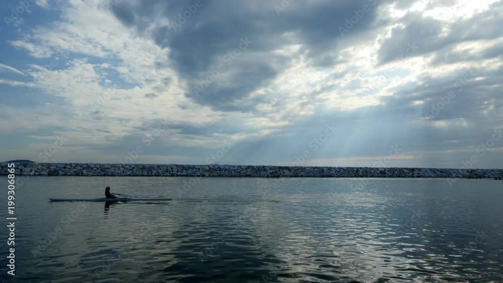 Young single scull rowing competitors paddle on the tranquil lake. Beautiful beams of the sunshine filter through clouds Young single scull rowing competitors paddle on the tranquil lake. Beautiful beams of the sunshine filter through clouds