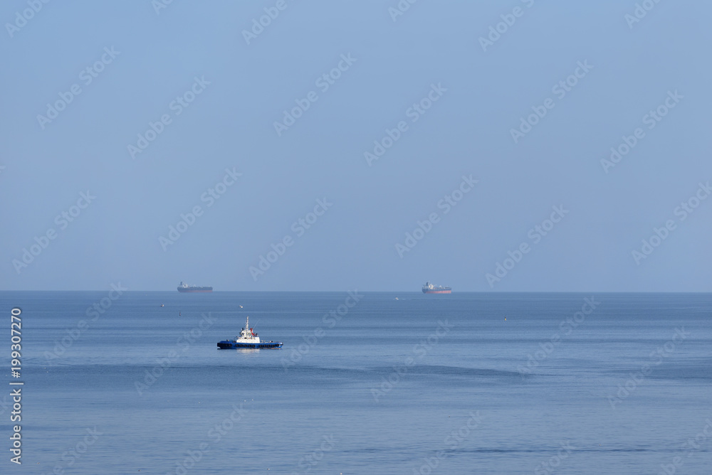 The tugboat stands without traffic in the sea. Tug ship alone in the sea against a blue sky background and large ships
