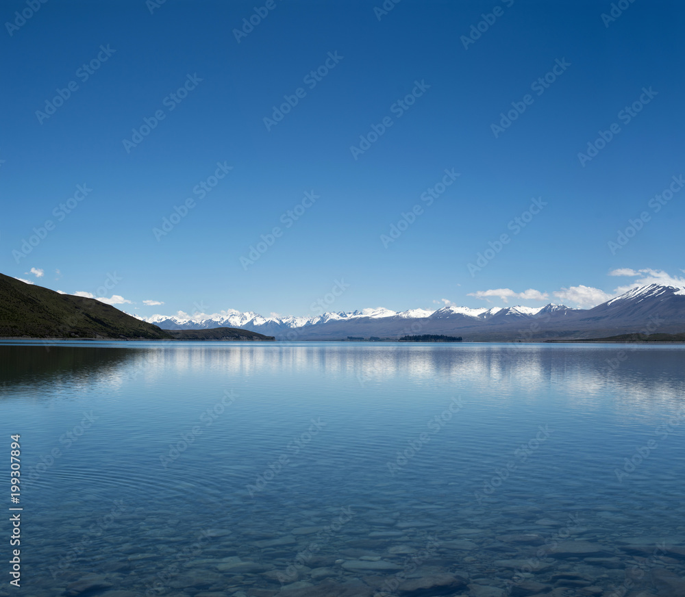 Imagen de un paisaje azul minimalista con montañas nevadas distantes y un lago enorme y claro. Las montañas se reflejan en el agua. Y hay muchas rocas y piedras en el fondo del lago.