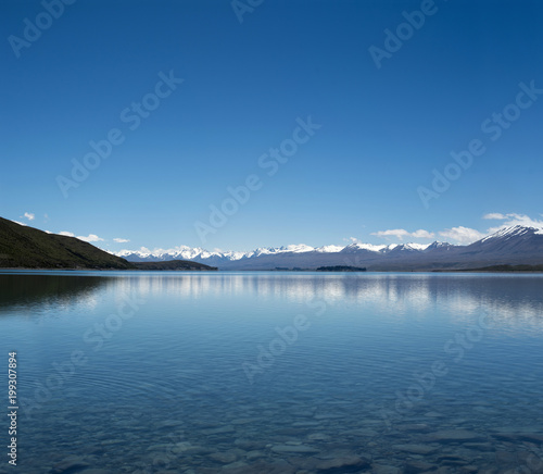 Imagen de un paisaje azul minimalista con monta  as nevadas distantes y un lago enorme y claro. Las monta  as se reflejan en el agua. Y hay muchas rocas y piedras en el fondo del lago.