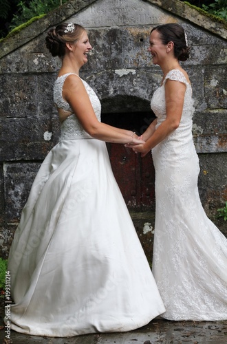 Two brides holding hands and gazing at each other in front of an ancient stone building , same sex wedding, both beautiful women in traditional white dresses 