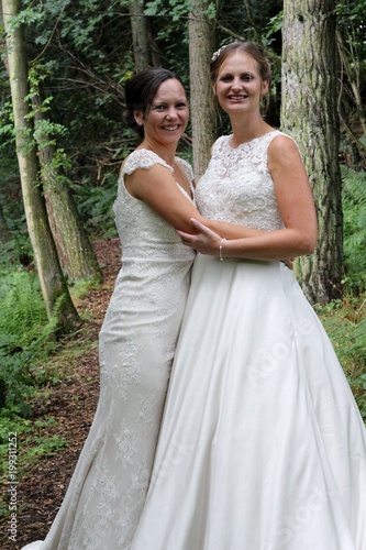 Brides in the woods, two smiling women on their same sex wedding day , both wearing traditional white dresses, posing in the trees