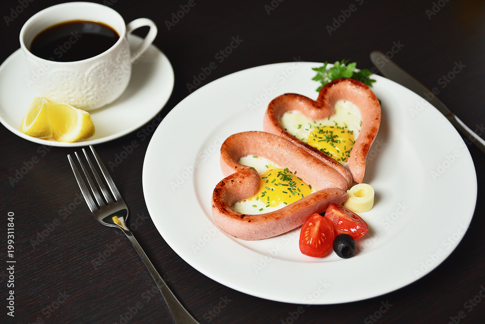 Scrambled eggs in the form of heart on a white plate with sausages, tomatoes, greens and coffee with lemon. Scrambled eggs in the form of heart on a white plate with sausages, tomatoes, greens and coffee with lemon.
