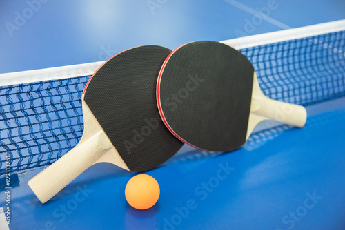 Orange ball for table tennis and two rackets of red and black color on a blue table with a grid