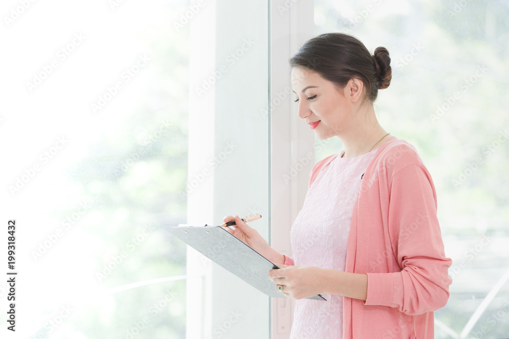 Business woman standing working near a window and holding clipboard at office. Business woman standing working near a window and holding clipboard at office.