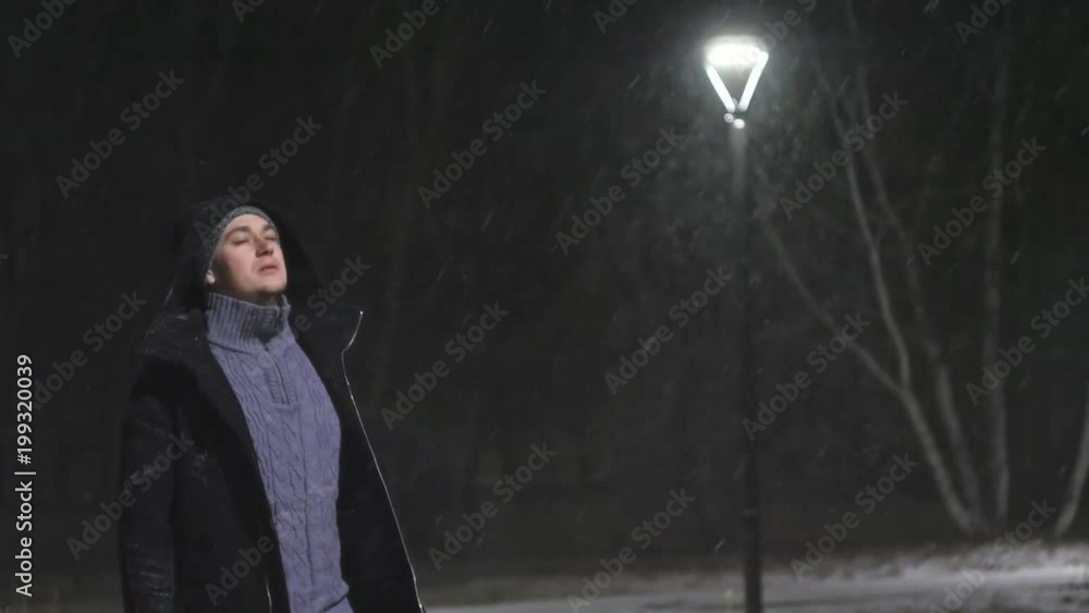The man in the street light. It's snowing against the background of lamp. Slow motion in 60fps. Snowflakes beautifully fall. The man is dressed in a cool black coat. The man in the street light. It's snowing against the background of lamp. Slow motion in 60fps. Snowflakes beautifully fall. The man is dressed in a cool black coat.