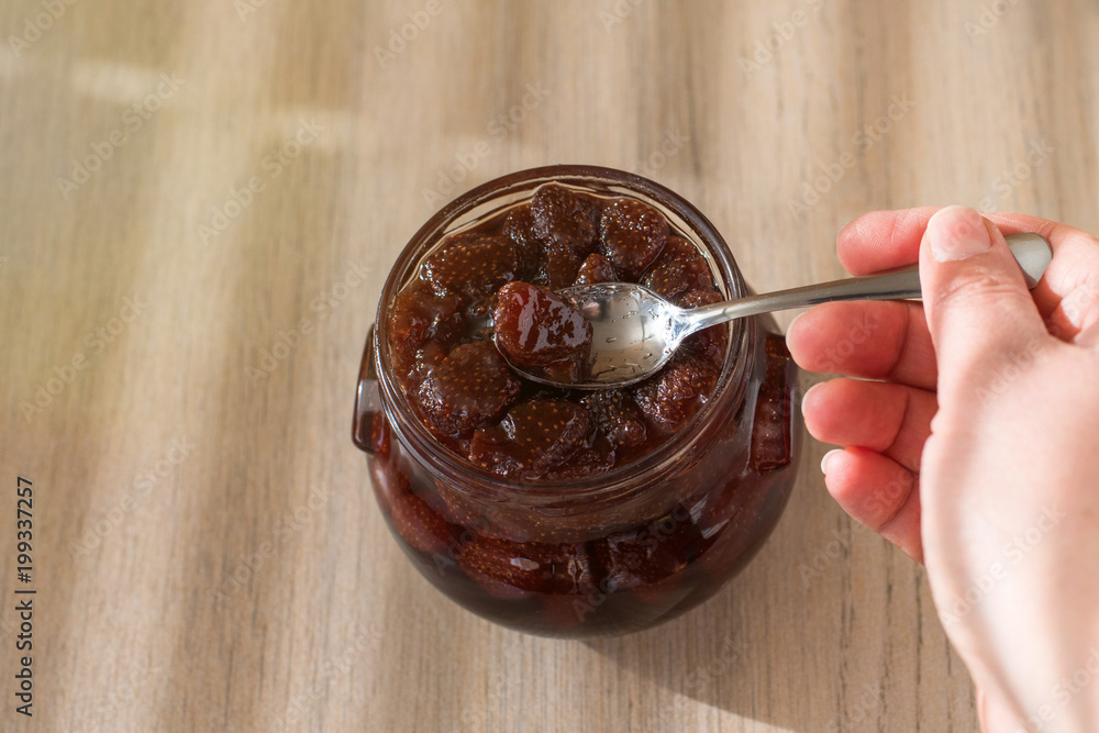 hand with a spoon and Jar of strawberry jam on light wooden background from top view