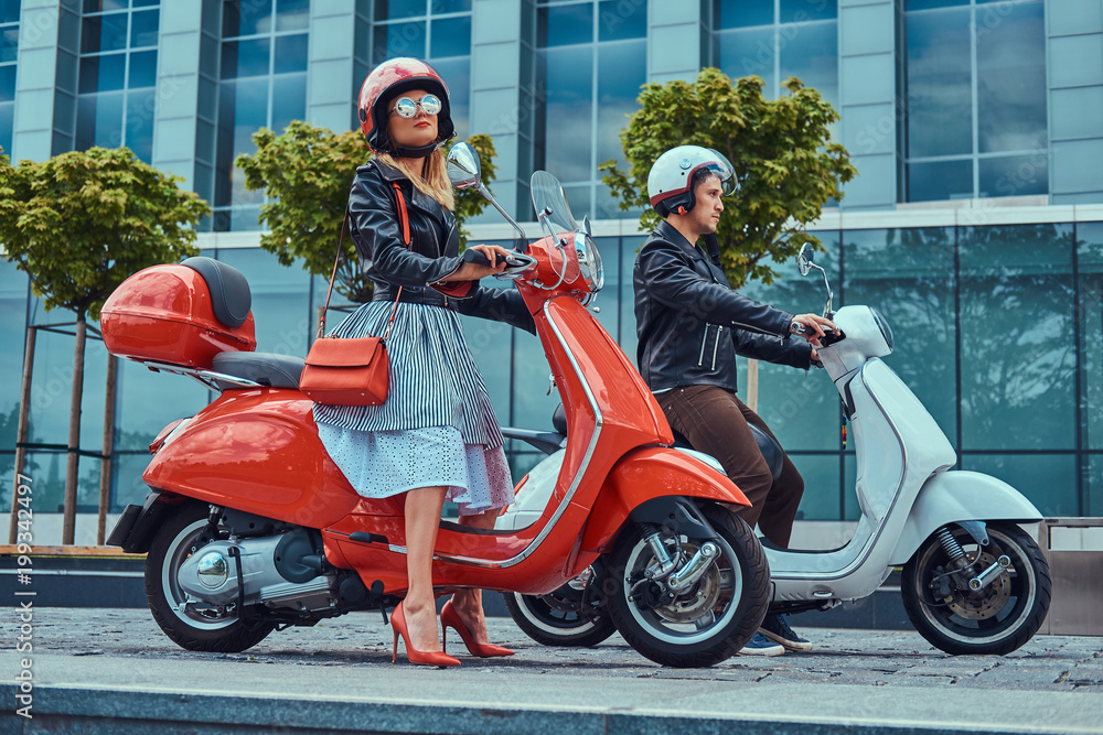 Attractive romantic couple, a handsome man and sexy female, sitting on retro Italian scooters against a skyscraper.