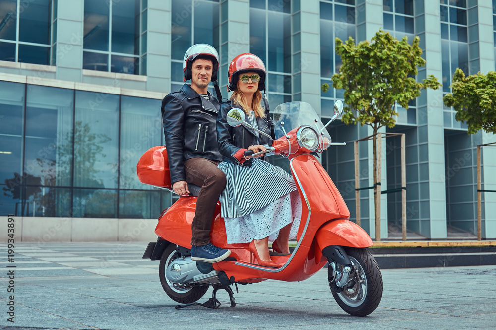 An attractive couple, a handsome man and sexy female riding together on a red retro scooter in a city.