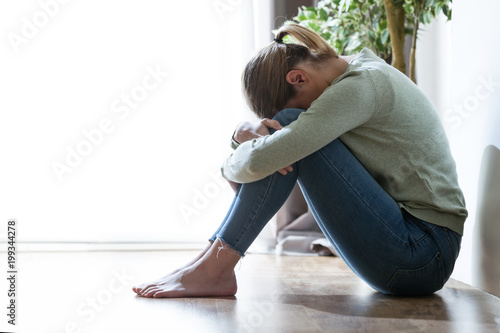 Unhappy lonely and depressed young woman hiding her face between legs at home.