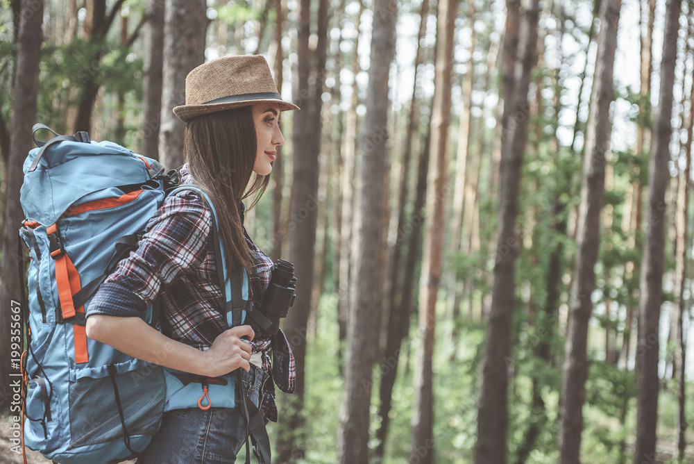 Action adventure. Side view profile of pleasant optimistic young woman wearing hat is standing in forest with rucksack and binoculars. She is looking ahead with slight smile. Copy space in right side