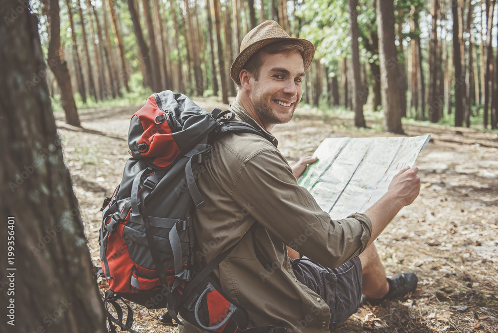 Joyful day. Side view of positive pleasant hiker with backpack is sitting on the ground in woodland and holding map. He is looking at camera with joy Joyful day. Side view of positive pleasant hiker with backpack is sitting on the ground in woodland and holding map. He is looking at camera with joy
