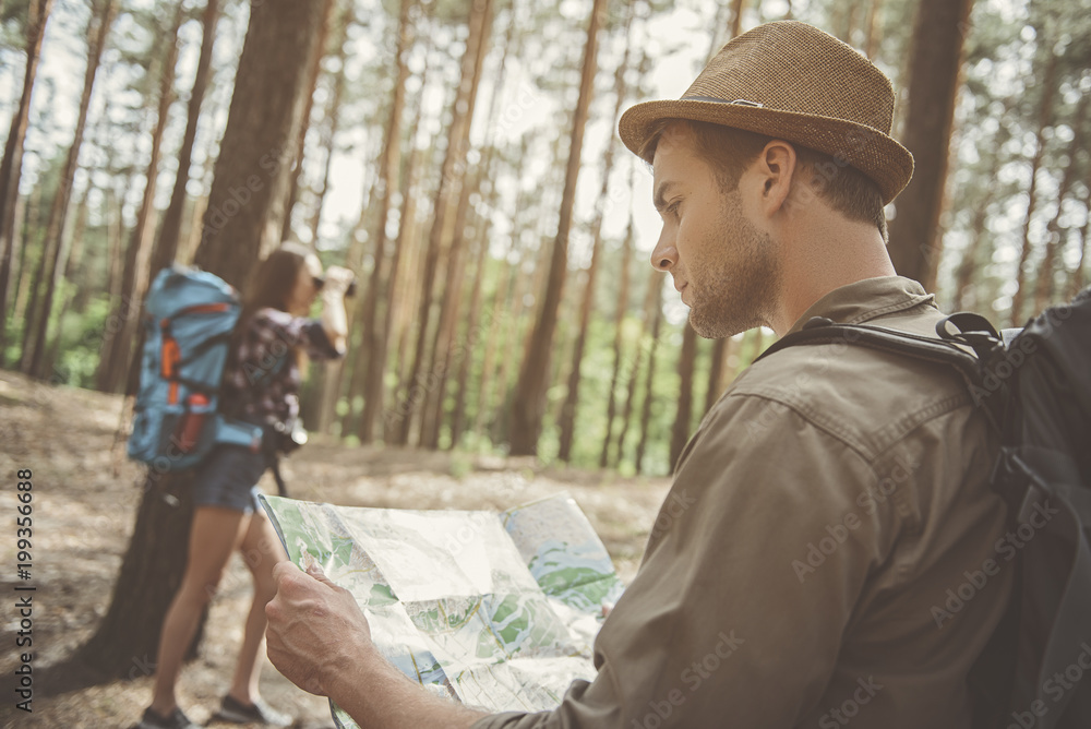 Where we are. Side view of concentrated young man is standing in the forest with his girlfriend. He is holding map while his girlfriend is looking into binoculars. Focus on man