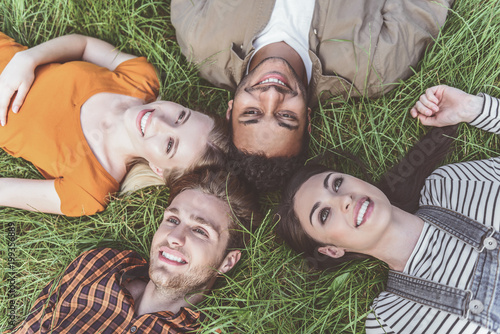 girls and guys lying on green grass with heads together. They are looking at the sky and smiling. Top view portrait