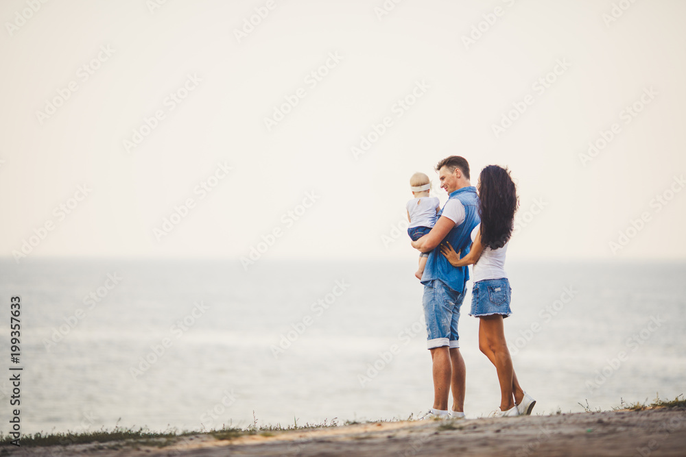 Family holiday in nature. Three, Mom, Dad, daughter one year standing with backs on cliff overlooking sea. man holding hild in arms,woman hugging husband.Fashionable people dressed in stylish clothes