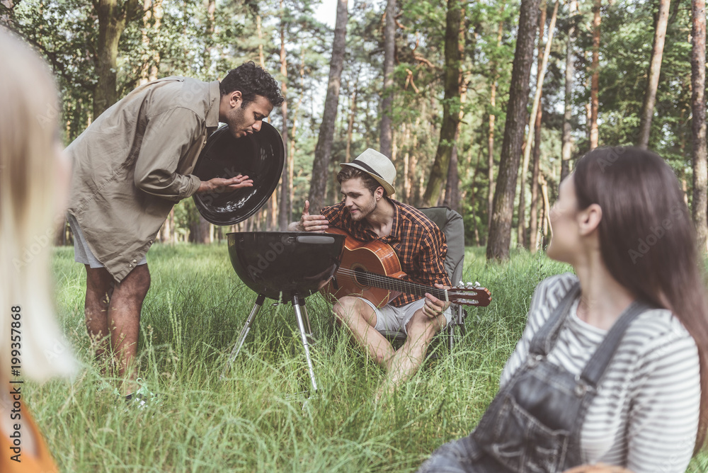 Fototapeta premium Two men opening barbecue cover and smelling smoke with pleasure. Smiling women sitting and looking at them