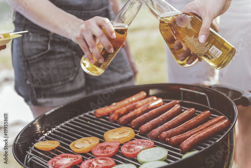 human hands clinking bottles of alcohol under grill. Vegetable and sausages are on grate. Close up