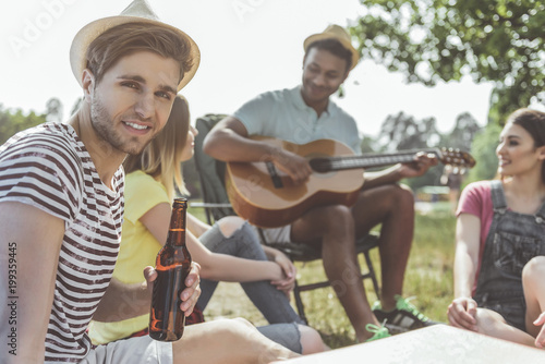 Two satisfied girls sitting on grass and listening to friend playing guitar. Focus on man holding bottle of alcohol and smiling