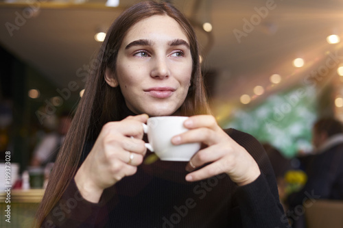 Young woman with long hair feel lonely, dreaming, drinking coffee in hands having rest in cafe near window