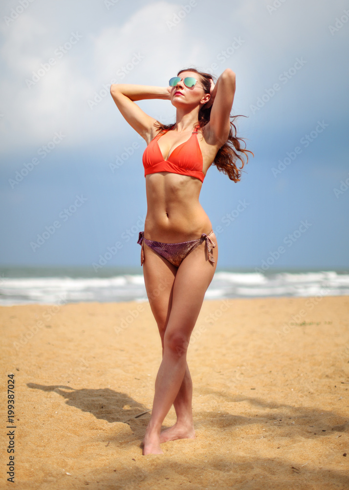 Young athletic woman in bikini and sunglasses, standing on the sand beach, holding her hand in hair behind her head, looking up, showing her flat stomach.