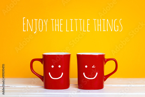 Two red coffee mugs with a smiling faces on a yellow background with the phrase Enjoy the little things.