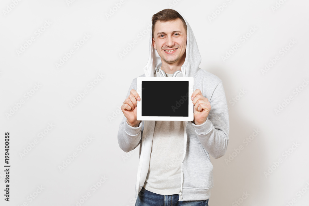Young handsome smiling man in t-shirt and light sweatshirt with hood with headphones holds tablet pc computer with blank screen for copy space isolated on white background. Concept of technology. Young handsome smiling man in t-shirt and light sweatshirt with hood with headphones holds tablet pc computer with blank screen for copy space isolated on white background. Concept of technology.