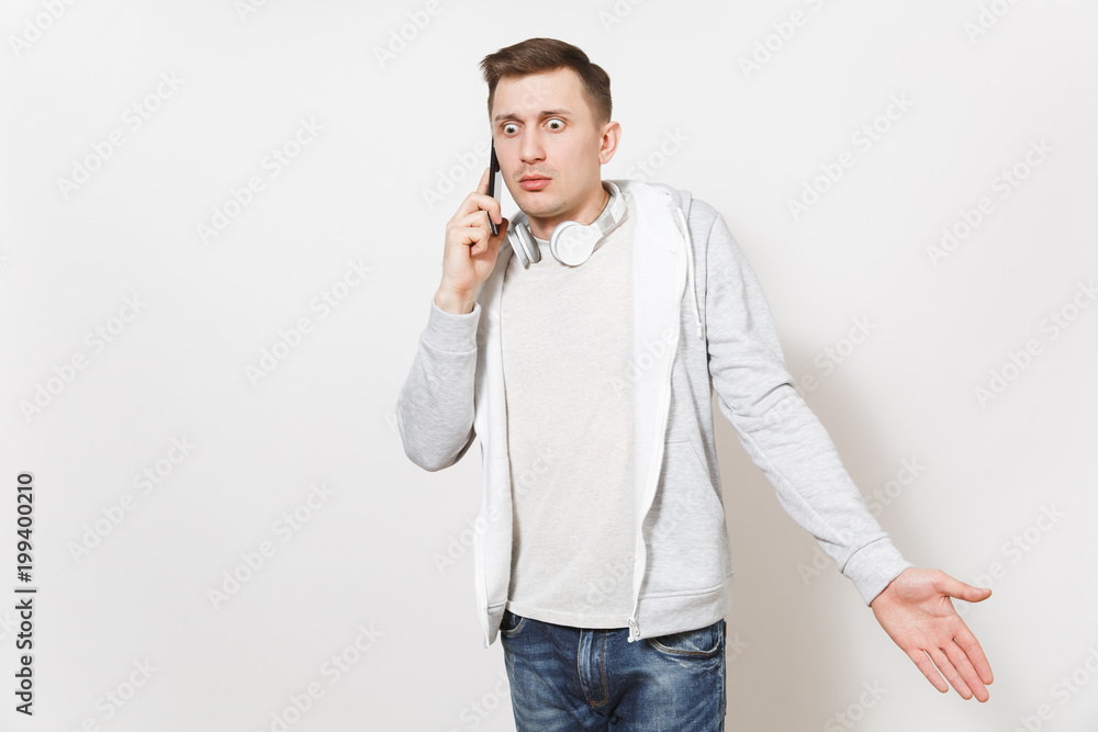 Young handsome troubled man student in t-shirt, light sweatshirt with headphones around neck talks on mobile phone and looks confused in studio on white background. Concept of communication, emotions. Young handsome troubled man student in t-shirt, light sweatshirt with headphones around neck talks on mobile phone and looks confused in studio on white background. Concept of communication, emotions.