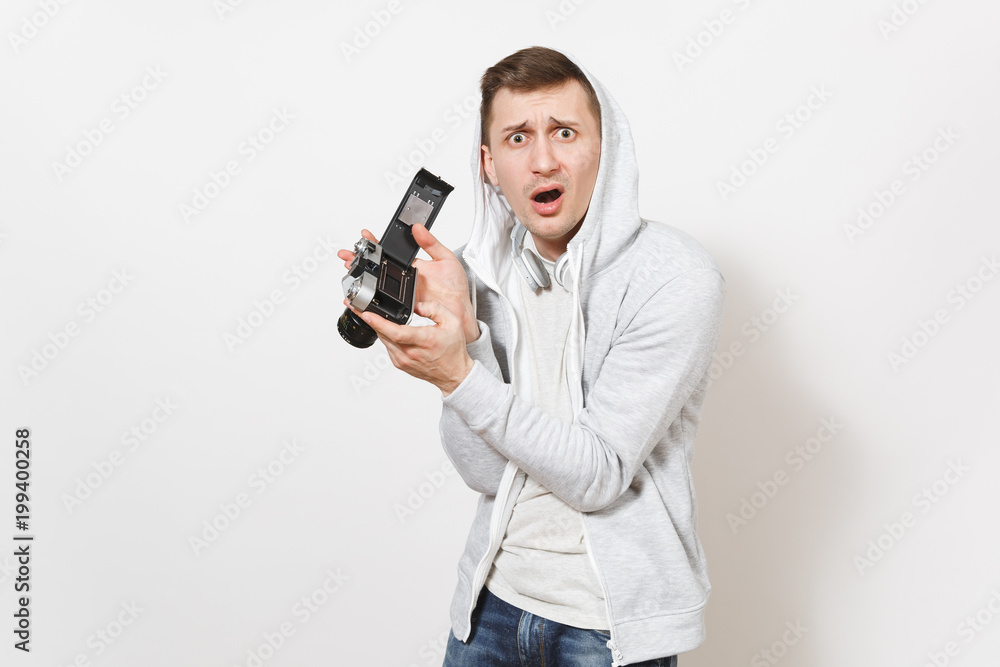 Young handsome surprised man student in t-shirt and light sweatshirt with hood with headphones holds opened retro camera and shows it to camera isolated on white background. Concept of photography Young handsome surprised man student in t-shirt and light sweatshirt with hood with headphones holds opened retro camera and shows it to camera isolated on white background. Concept of photography