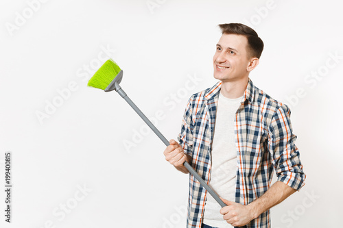 Young smiling happy housekeeper man in checkered shirt holding and sweeping with green broom isolated on white background. Male doing house chores. Copy space for advertisement. Cleanliness concept.