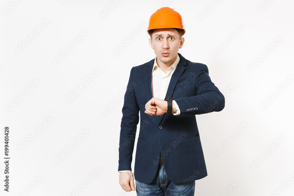 Young concerned dissatisfied businessman in dark suit, protective construction orange helmet with clock on hand isolated on white background. Time is running out. Male worker for advertisement. Young concerned dissatisfied businessman in dark suit, protective construction orange helmet with clock on hand isolated on white background. Time is running out. Male worker for advertisement.