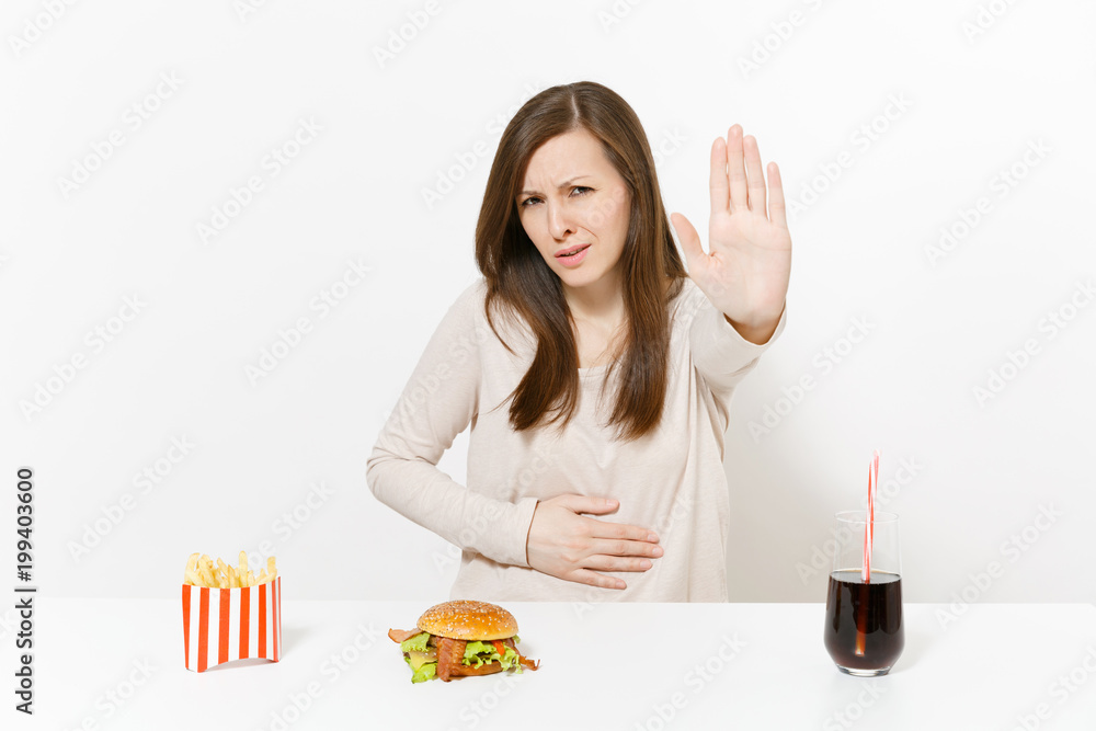 Illness woman put hand on pain abdomen, stomach-ache at table with burger, french fries, cola in glass bottle isolated on white background. Proper nutrition or American classic fast food. Copy space. Illness woman put hand on pain abdomen, stomach-ache at table with burger, french fries, cola in glass bottle isolated on white background. Proper nutrition or American classic fast food. Copy space.