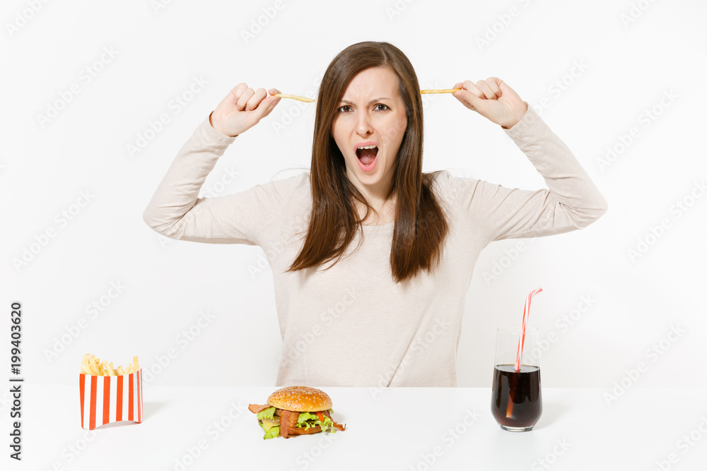 Dissatisfied woman at table with several pieces of potatoes, french fries, burger, cola in bottle isolated on white background. Proper nutrition or American classic fast food. Area with copy space. Dissatisfied woman at table with several pieces of potatoes, french fries, burger, cola in bottle isolated on white background. Proper nutrition or American classic fast food. Area with copy space.