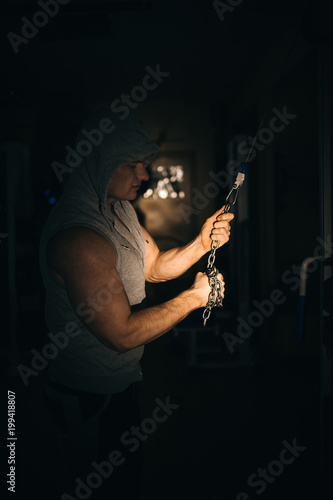 an athlete pumps his arm muscles with a chain in the gym on a dark background,