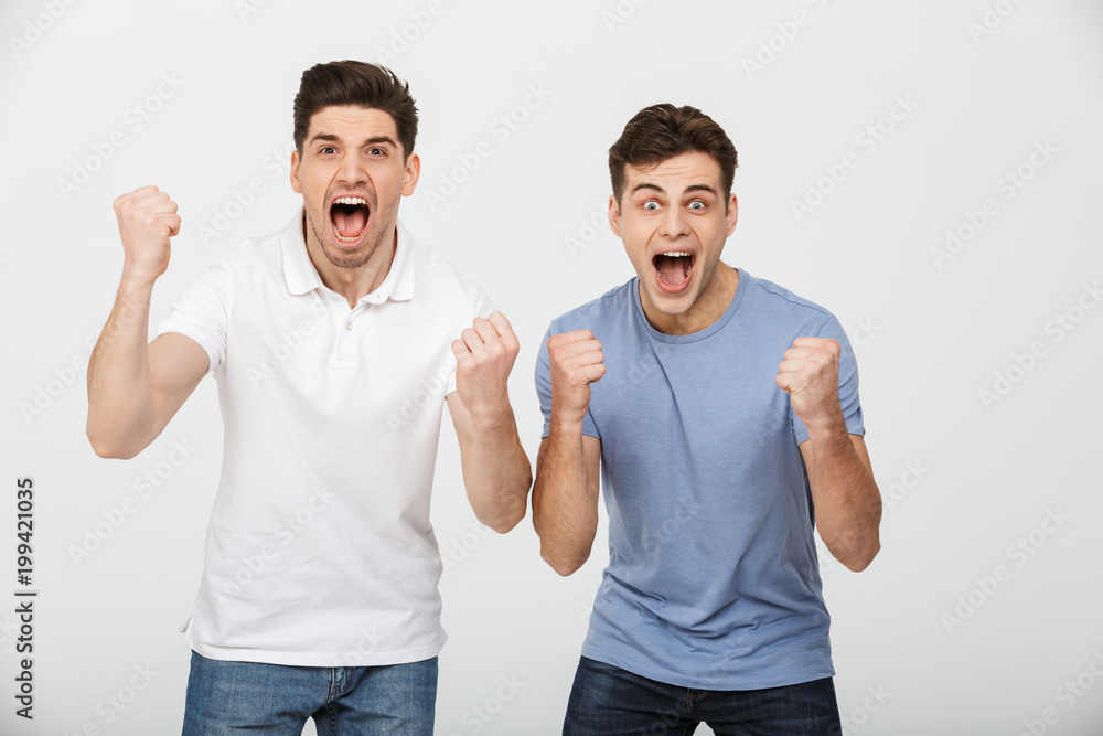 Photo of two ecstatic men 30s wearing casual t-shirt and jeans shouting and rejoicing victory with clenching fists, isolated over white background Photo of two ecstatic men 30s wearing casual t-shirt and jeans shouting and rejoicing victory with clenching fists, isolated over white background