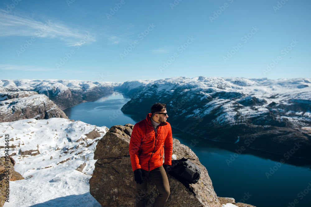 Man tourist in sunglasses seating on a stone and looking at river at lysefjord in norway, beautiful sunny winter weather