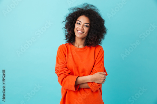 Colorful portrait of amazing woman in red shirt with afro hairstyle looking on camera with smile, isolated over blue background