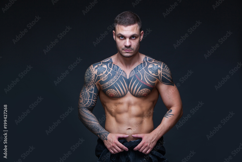 The man with a muscular naked torso with tattoos on the dark background. ABS The man with a muscular naked torso with tattoos on the dark background. ABS