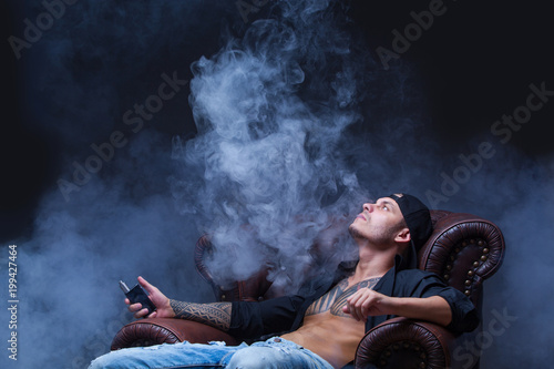 Vaper. The man with tattoos sits on a leather sofa smoke an electronic cigarette on the dark background.