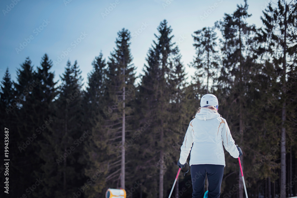 Female skier in white jacket and protective helmet on ski slope with pine trees. Rear view.