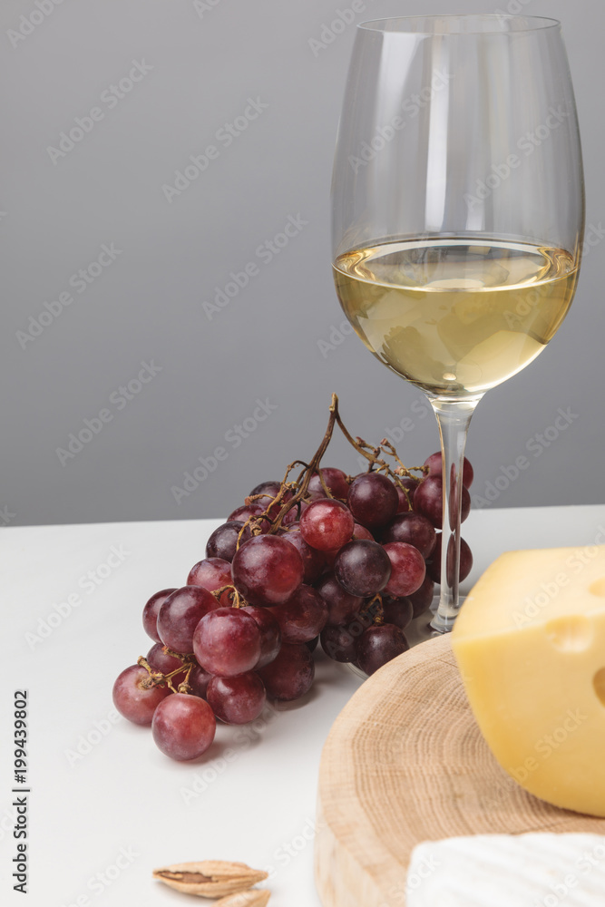 Partial view of maasdam cheese on wooden board, wine glass, almond and grapes on gray