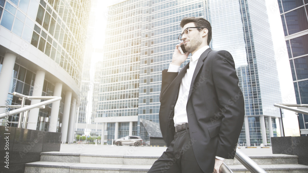 A smiling young businessman dressed in a black suit is talking on the phone on the stairs. Sunlight, downtown and city on the background A smiling young businessman dressed in a black suit is talking on the phone on the stairs. Sunlight, downtown and city on the background