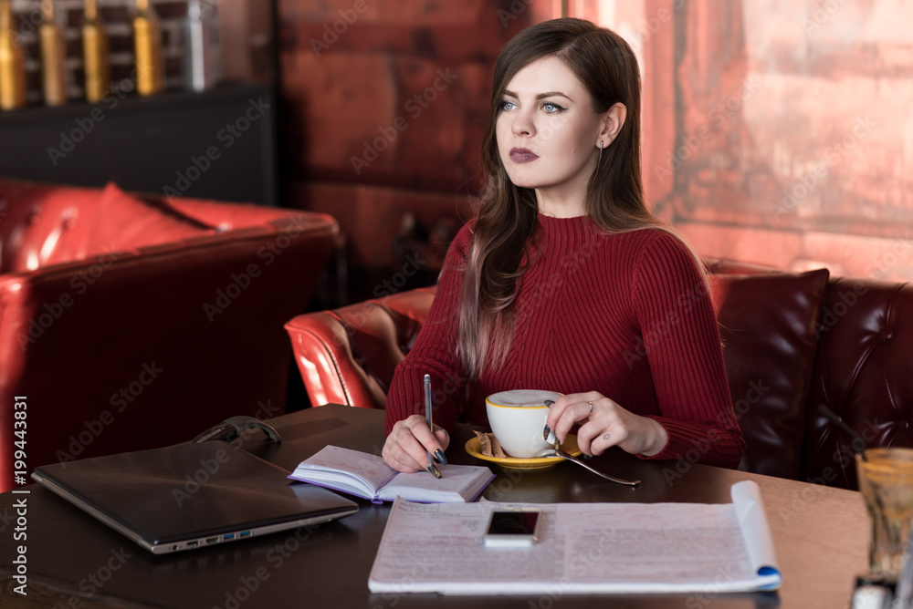 Beautiful business woman writes a to-do list in a notebook. Serious view. A cup of coffee in hand. Laptop and phone on the table. Red Turtleneck. Leather sofa.