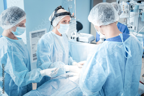 Surgical team. Professional smart medical assistant wearing uniform and holding an instrument while doing her job