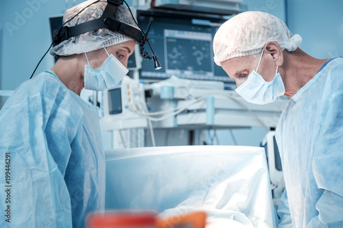 Medical workers. Serious smart professional surgeons standing opposite each other and looking at the patient while working