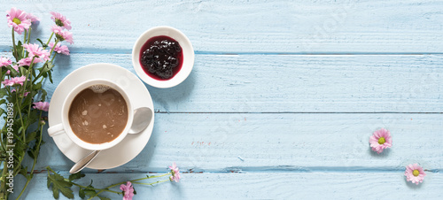 Coffee cup, jam and pink flowers on a pastel blue wooden background with copy space, top view from above, panoramic banner format
