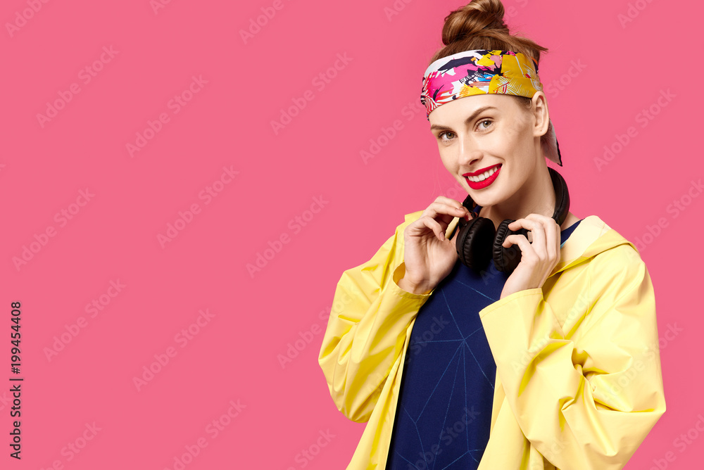 Young beautiful woman on a pink background and a yellow coat listening to music through wireless headphones. Concept of healthy nutrition and sports. Colour obsession concept. Young beautiful woman on a pink background and a yellow coat listening to music through wireless headphones. Concept of healthy nutrition and sports. Colour obsession concept.