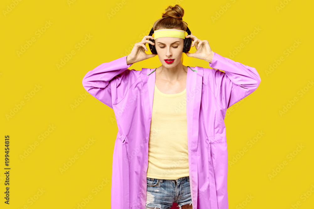 Young woman on a yellow background and a pink coat hugs music worm wireless headphones. Colour obsession concept. Minimalistic style. Stylish Trendy Young woman on a yellow background and a pink coat hugs music worm wireless headphones. Colour obsession concept. Minimalistic style. Stylish Trendy