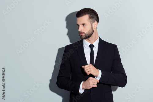 Portrait of cool, virile, harsh guy with half-turned face to the side fasten button on cuff link of his white shirt, going for date with lover, isolated on grey background